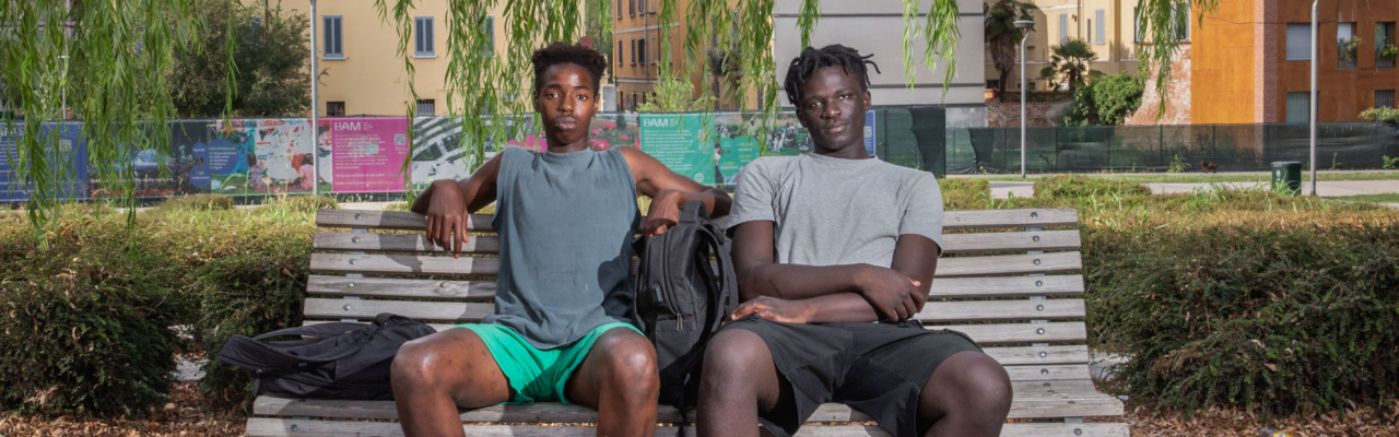 Ndongo Gueye, 18, and Cheikh Badar Gueye, 19, born in Senegal and raised in northern Italy, sit in the shade at the BAM Tree Library, a park in the financial district of Milan. Italy, 2022 - Humidity 38%, Temperature 35°C